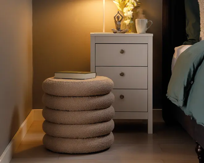 En rund, blød puf med et unikt design bestående af fire stablede ringe, betrukket med et blødt, beige tekstil med tekstur. Puffen har tykke, afrundede lag, der giver et moderne og hyggeligt udtryk. Øverst ligger en lukket bog, hvilket viser, at puffen også kan bruges som et lille bord eller ekstra siddeplads.