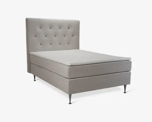 En moderne seng med en høj, polstret sengegavl med knap-tuftet design. Sengens ramme og madrasbase er betrukket med et lysebeige stof, hvilket giver et rent og elegant udseende. Sengen står på slanke, metalliske ben, som tilføjer et moderne præg. Det samlede design er minimalistisk med lige linjer og et blødt, polstret look for komfort.