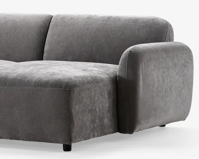 Billedet viser en del af en moderne sofa med blødt, velouragtigt gråt stof. Sofaen har en kraftig, afrundet armlæn og en bred, dyb siddepude, som giver god komfort. Ryglænet er let buet, og designet er minimalistisk med glatte, rene linjer. Sofaen står på korte, solide ben, som næsten ikke kan ses.