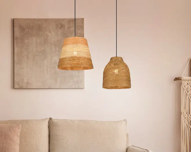 To hængelamper med håndvævede lampeskærme af naturfibre hænger fra loftet. Lampen til venstre har en konisk form med skiftevis lyse og mellembrune bånd, mens lampen til højre har en rund, klokkeformet skærm i ensartet lysebrun farve. Begge skærme har en tekstureret, rustik overflade, og ledningerne er tynde og sorte. Lamperne udsender et varmt, blødt lys, der understreger rummets hyggelige og indbydende stemning.