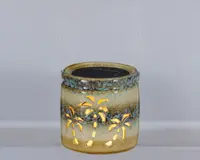 Keramisk lanterne med blank, flerfarvet glasur i beige, brun og turkis nuancer. Lanternen har udskårne palmetræ-motiver, som lyset kan skinne igennem og skabe et dekorativt mønster. Den er rund i formen og har et metalhåndtag, så den er nem at bære eller hænge op. Øverst ses et solcellepanel, hvilket tyder på, at den er solcelledrevet.