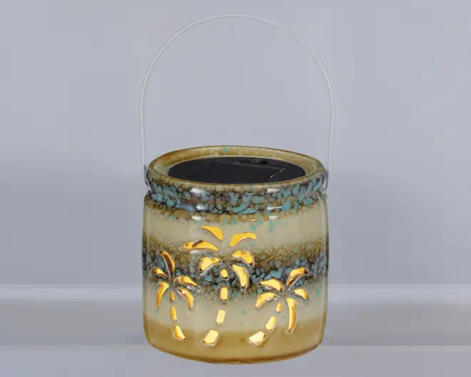 Keramisk lanterne med blank, flerfarvet glasur i beige, brun og turkis nuancer. Lanternen har udskårne palmetræ-motiver, som lyset kan skinne igennem og skabe et dekorativt mønster. Den er rund i formen og har et metalhåndtag, så den er nem at bære eller hænge op. Øverst ses et solcellepanel, hvilket tyder på, at den er solcelledrevet.