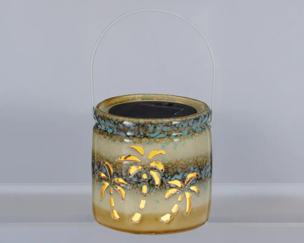 Keramisk lanterne med blank, flerfarvet glasur i beige, brun og turkis nuancer. Lanternen har udskårne palmetræ-motiver, som lyset kan skinne igennem og skabe et dekorativt mønster. Den er rund i formen og har et metalhåndtag, så den er nem at bære eller hænge op. Øverst ses et solcellepanel, hvilket tyder på, at den er solcelledrevet.