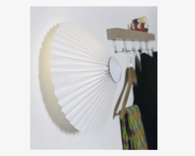 Væghængt lampe med en plisseret hvid skærm, der folder sig ud fra en central cirkulær base og skaber en blød, diffus lyseffekt på væggen. Lampen har et moderne, minimalistisk design og er placeret tæt på en knagerække med hængende tøj og tilbehør.