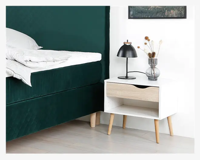 Et moderne natbord med en hvid, rektangulær krop, en enkelt skuffe i lyst træ med et buet udskåret greb og fire skråtstillede ben i lyst træ. Bordet har en åben hylde under skuffen. Ovenpå står en sort metalbordlampe med kuppelformet skærm og en glaskrukke med tørrede blomster.