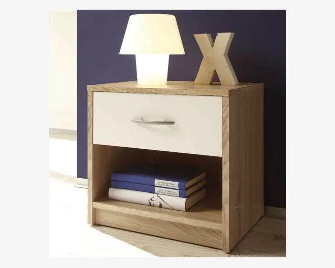 Et moderne natbord med lys egetræsfinish og en enkelt hvid skuffe med et slankt sølvhåndtag. Under skuffen er der en åben hylde med tre stablede bøger. På toppen står en tændt bordlampe med hvid skærm samt et dekorativt bogstav 'X' i træ. Designet er minimalistisk og funktionelt, velegnet til moderne soveværelser.