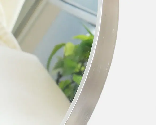Nærbillede af et rundt spejl med en elegant, børstet metalramme. Spejlet reflekterer et lyst rum med et vindue og nogle grønne planter, hvilket antyder et moderne og luftigt interiør. Rammen har en glat overflade og en diskret metallisk glans.
