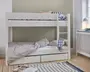 Hvid køjeseng i træ med et minimalistisk design, bestående af to enkeltsenge stablet oven på hinanden. Den nederste seng har stribet dyne og pude, mens den øverste har puder med geometrisk mønster. En solid stige er fastgjort i højre side for nem adgang til overkøjen. Under den nederste seng er der to rummelige udtræksskuffer til ekstra opbevaring. Sengerammen har rene linjer og et moderne udtryk, velegnet til børne- eller gæsteværelse.
