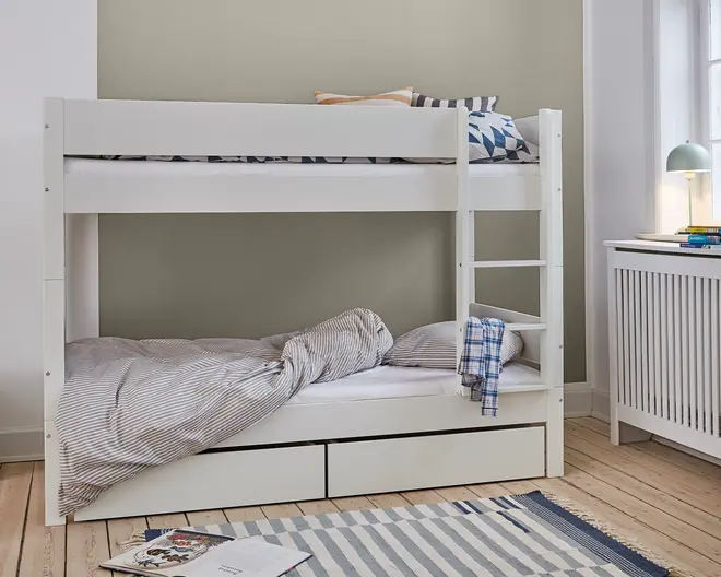 Hvid køjeseng i træ med et minimalistisk design, bestående af to enkeltsenge stablet oven på hinanden. Den nederste seng har stribet dyne og pude, mens den øverste har puder med geometrisk mønster. En solid stige er fastgjort i højre side for nem adgang til overkøjen. Under den nederste seng er der to rummelige udtræksskuffer til ekstra opbevaring. Sengerammen har rene linjer og et moderne udtryk, velegnet til børne- eller gæsteværelse.