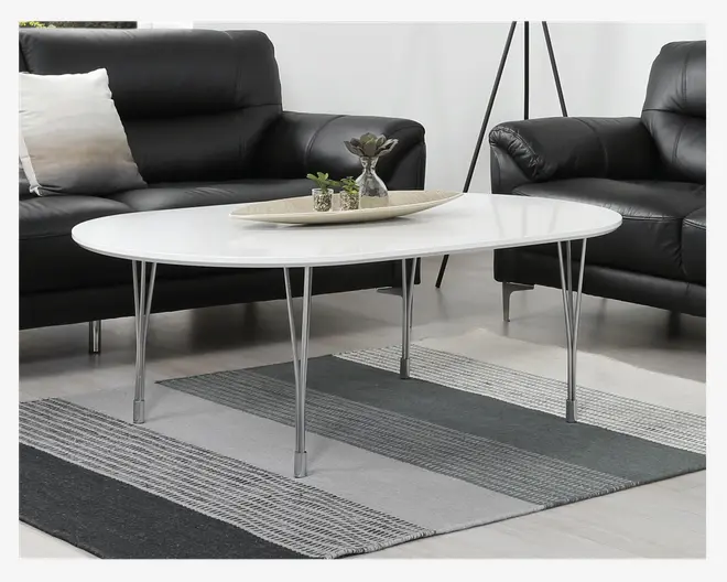 Ovalt bord med blank hvid bordplade og slanke, skråtstillede ben i krom. Bordet har et minimalistisk design og står på et stribet tæppe. Ovenpå står et rektangulært fad med små dekorative planter og en vase.