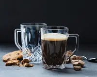 To klare glas krus med solide håndtag og et facetslebet diamantmønster i bunden. Det ene krus er fyldt med en mørk drik med et lag skum på toppen, mens det andet er tomt. Krusene er omgivet af brune sukkerknalder, en metalske og chokoladechip cookies, hvoraf nogle er brækket i stykker.