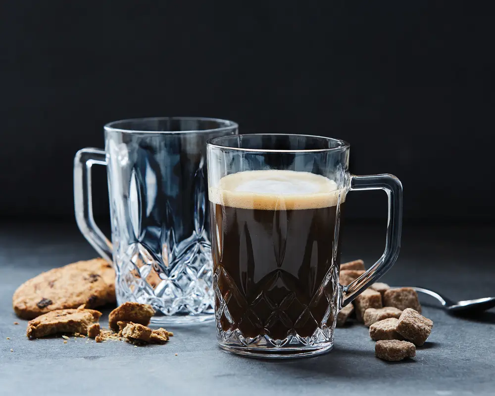 To klare glas krus med solide håndtag og et facetslebet diamantmønster i bunden. Det ene krus er fyldt med en mørk drik med et lag skum på toppen, mens det andet er tomt. Krusene er omgivet af brune sukkerknalder, en metalske og chokoladechip cookies, hvoraf nogle er brækket i stykker.