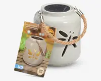 Hvid keramisk lanterne med udskåret guldsmed-design på siden, udstyret med solcellepanel på toppen til opladning. Lanternen har et kraftigt, flettet rebhåndtag fastgjort med metalbeslag, så den nemt kan bæres eller hænges op. Den udsender varmt hvidt LED-lys og er velegnet til udendørs brug.