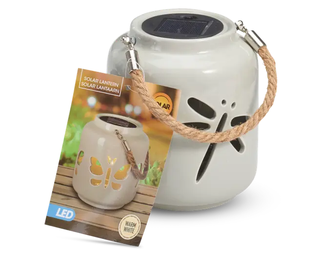 Hvid keramisk lanterne med udskåret guldsmed-design på siden, udstyret med solcellepanel på toppen til opladning. Lanternen har et kraftigt, flettet rebhåndtag fastgjort med metalbeslag, så den nemt kan bæres eller hænges op. Den udsender varmt hvidt LED-lys og er velegnet til udendørs brug.