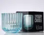 En transparent, lyseblå glaslysestage med lodret rillet struktur. Lysestagen er cylindrisk med en let udvidet bund og vises med flere små tændte lys indeni. Glasset fremstår tykt og robust med et moderne, elegant design. Emballagen i baggrunden viser et billede af lysestagen og angiver en højde på 6,5 cm.
