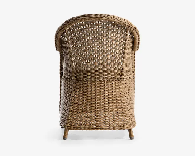 Billedet viser bagsiden af en stol fremstillet af tæt flettet natur-rattan. Stolen har en rund, tøndeformet ryg og armlæn, der buer elegant ned mod sædet. Fletmønstret er ensartet og tæt, hvilket giver en solid og støttende konstruktion. Benene er korte og let skrå, også dækket af rattan for et ensartet udseende. Overfladen har en varm, lys brun farve, der fremhæver materialets naturlige struktur.