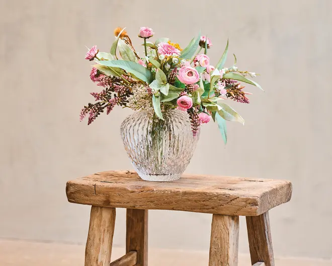 En klar, rillet glasvase med rund form, fyldt med en blandet buket af lyserøde og hvide blomster, grønt løv og nogle tørrede elementer. Buketten indeholder forskellige blomsterarter, herunder små runde blomster og aflange blade, hvilket giver et naturligt og let vildt udtryk. Vasens placeres på en rustik træskammel med en rå, ubehandlet overflade.