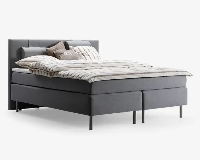 En moderne seng med gråt polstret stel og hovedgærde, udstyret med slanke metalben. Sengen er redt med lyst, stribet sengetøj og har flere pyntepuder i neutrale nuancer, hvilket giver et hyggeligt og indbydende udtryk. Designet er minimalistisk og moderne.