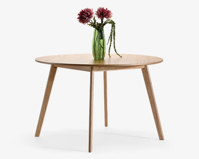 Rundt bord i lyst træ med glat bordplade og fire tilspidsede skrå ben, dekoreret med en grøn struktureret vase med lyserøde blomster og hængende grønt.