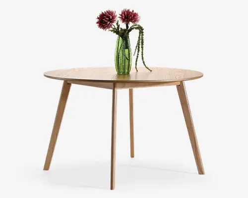 Rundt bord i lyst træ med glat bordplade og fire tilspidsede skrå ben, dekoreret med en grøn struktureret vase med lyserøde blomster og hængende grønt.