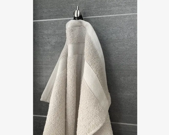 Et lysebeige håndklæde med blød, fyldig tekstur og en dekorativ vævet kant. Håndklædet ser tykt og absorberende ud, og kanten har et diskret mønster for ekstra stil. Det hænger på en krog foran en grå flisevæg.