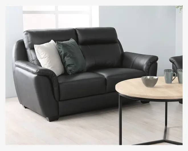 En moderne to-personers sofa betrukket med glat sort læder, med bløde, polstrede armlæn og høje ryglæn for ekstra komfort. Sofaen har to pyntepuder, én i hvid og én i mørkegrøn, som giver et stilfuldt kontrast. Designet er elegant med let buede armlæn og står på korte, solide ben.
