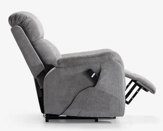 Sidebillede af en moderne lænestol betrukket med lysegråt stof. Stolen er fuldt tilbagelænet med udslået fodstøtte samt en tykt polstret ryg- og siddeflade for ekstra komfort. Der er en ledningsforbundet fjernbetjening fastgjort på siden, hvilket indikerer elektriske justeringsfunktioner. Armlænene er brede og polstrede, og designet er slankt og moderne.