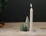 Et hvidt stearinlys står oprejst i en keramisk stage, som er en del af en dekorativ opstilling med tre små, stiliserede keramiske træer i forskellige nuancer af grøn og grå. Træerne er kegleformede og har en blank overflade, hvoraf to ligner klassiske juletræer, og det tredje er en glat kegle. Opstillingen står på en rund, hvid base, og der er kunstig sne drysset omkring, hvilket skaber en festlig og vinterlig stemning.