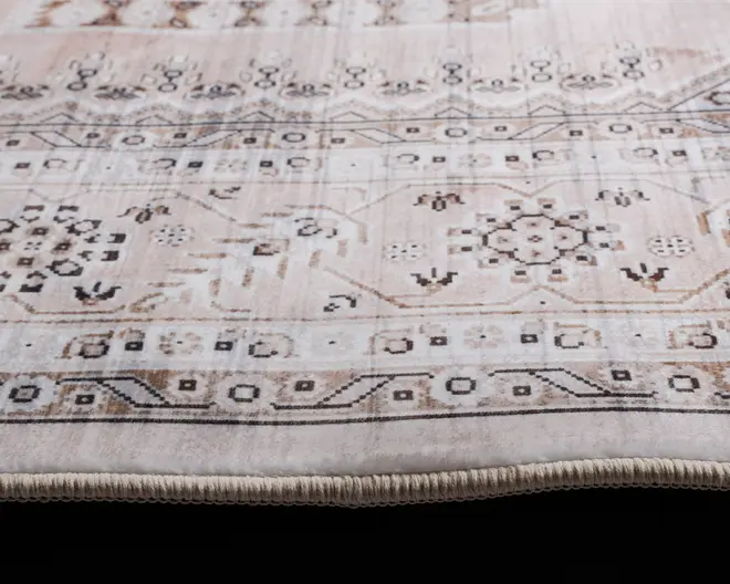 Nærbillede af et tæppe med en blød, neutral farvepalet i beige, creme og lysebrune nuancer. Tæppet har detaljerede geometriske og blomsteragtige mønstre, herunder sekskantede former og stiliserede motiver, arrangeret i vandrette bånd. Kanten er afsluttet med tæt vævet kantbånd, hvilket sikrer holdbarhed og et pænt udseende. Overfladen fremstår blød og glat, ideel til at tilføje et diskret dekorativt præg til forskellige interiører.