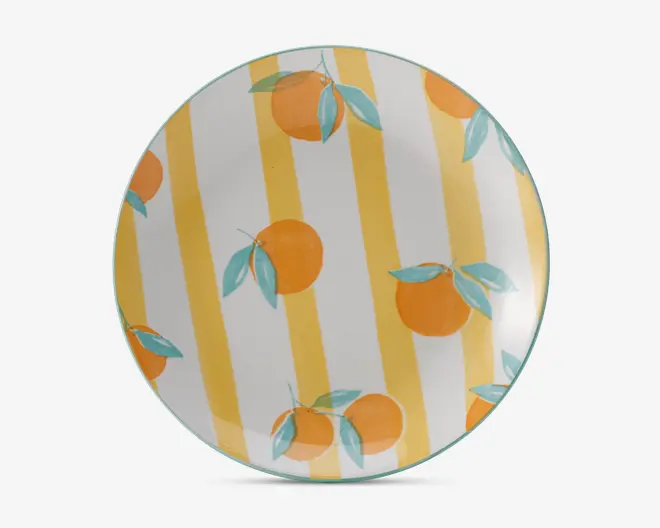 Rund tallerken med et legende design, der viser håndmalede appelsiner og grønne blade spredt over overfladen. Baggrunden har brede, lodrette gule og hvide striber, hvilket giver et lyst og muntert udtryk. Kanten er diskret markeret med en lyseblå farve.