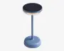 En moderne, cylindrisk bordlampe med mat blå finish. Lampen har en rund, flad top med en sort, tekstureret overflade, sandsynligvis en berøringsfølsom eller solpanel. Der er en gennemsigtig ring under toppen, som fungerer som lyskilde. Lampen har en slank, lige stang og en bred, stabil fod med et diskret tænd/sluk-symbol.