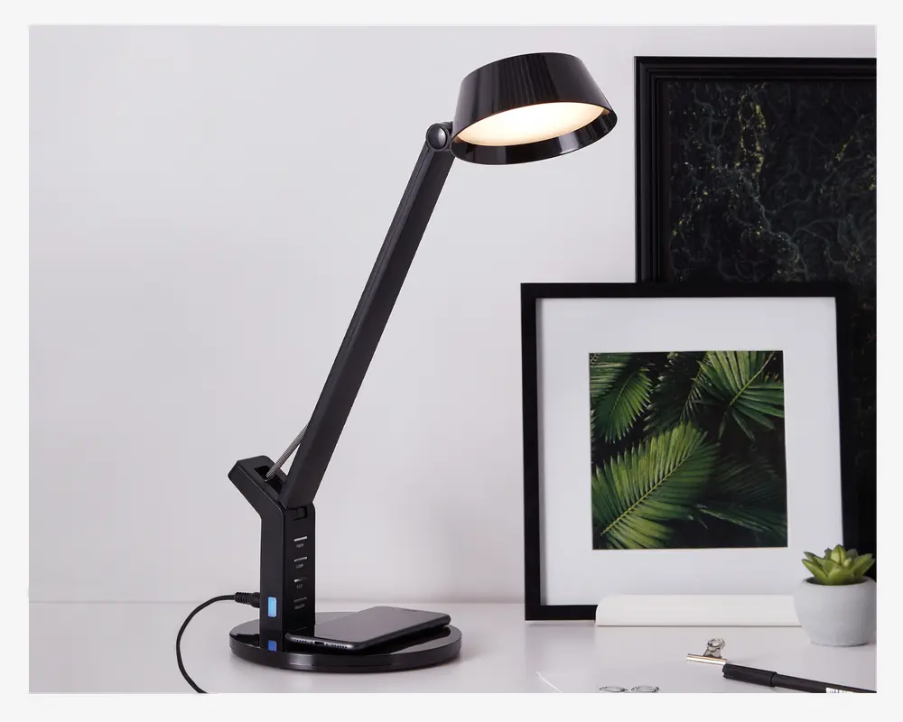 Elegant sort skrivebordslampe med justerbar arm og rund lampeskærm, udstyret med touch-kontrolpanel på basen. Basen har flere USB-opladningsporte og en trådløs opladningsplade til enheder. Lampen har et moderne, minimalistisk design, der passer til både kontor og hjem.