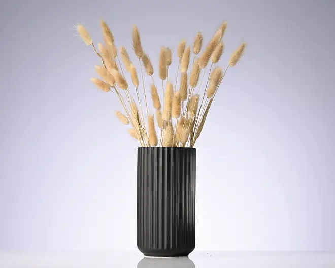 En høj sort cylindrisk vase med lodret rillet struktur, fyldt med en buket tørrede beige græsstrå med bløde, fjeragtige aks.