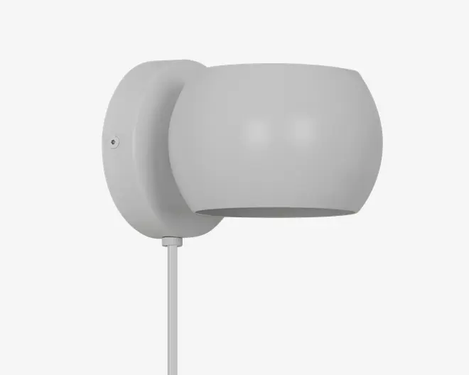 En moderne vægmonteret lampe med en glat, mat grå overflade. Lampen har en rund, cylindrisk skærm, der leder lyset nedad. Basen er cirkulær og matcher skærmen i farve og tekstur. En synlig ledning går ud fra bunden af lampen til strømtilslutning.