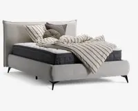 Polstret sengegavl med høj, blød hovedgavl i lysebeige stof med diskret geometrisk mønster. Sengen har en tyk, mørkegrå madras og en hvid topmadras. Sengetøjet består af en beige og gråstribet dyne, der ligger løst hen over sengen, samt flere pyntepuder i forskellige former og teksturer, herunder en rund, plisseret pude og firkantede puder i beige og mørkegrå. Sengen står på korte, skrå sorte ben.