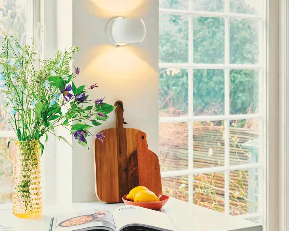 To skærebrætter af træ med naturlige træmønstre og afrundede håndtag står op ad en hvid væg. Det ene bræt er lidt større og mørkere end det andet. Begge brætter har en glat overflade og et hul i håndtaget til ophængning. De står på en hvid bordplade ved siden af en skål med citroner, en guldfarvet vase med grønne og lilla blomster samt en åben kogebog. Scenen er oplyst af naturligt lys fra et stort vindue.