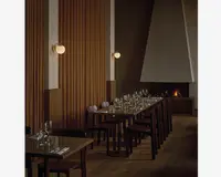 Billedet viser et moderne spiseområde med træborde og mørke træstole, der er arrangeret i pæne rækker. Hvert bord er dækket med klart glas, herunder vinglas og vandglas, samt hvide servietter og bestik. Rummet har varm belysning fra runde væglamper med messingbeslag, og en stor, moderne pejs med en geometrisk hvid hætte ses i baggrunden, hvilket skaber en hyggelig stemning. Væggene har lodrette træpaneler, og gulvet er af lyst træ, hvilket bidrager til den elegante og indbydende atmosfære.