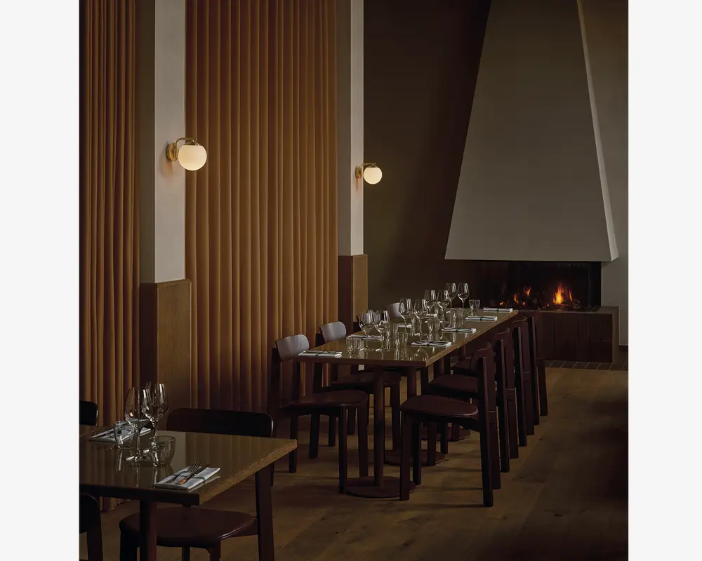 Billedet viser et moderne spiseområde med træborde og mørke træstole, der er arrangeret i pæne rækker. Hvert bord er dækket med klart glas, herunder vinglas og vandglas, samt hvide servietter og bestik. Rummet har varm belysning fra runde væglamper med messingbeslag, og en stor, moderne pejs med en geometrisk hvid hætte ses i baggrunden, hvilket skaber en hyggelig stemning. Væggene har lodrette træpaneler, og gulvet er af lyst træ, hvilket bidrager til den elegante og indbydende atmosfære.