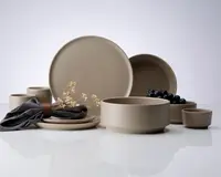 Et sæt mat beige keramikservice bestående af to store tallerkener, to mellemstore skåle, to små kopper og en lille skål. Delene har et minimalistisk, moderne design med glatte overflader og lige kanter. Sættet er stylet med en mørkegrå stofserviet holdt af en brun læderring og en kvist tørrede blomster, hvilket giver et elegant udtryk. Nogle af skålene indeholder blåbær, hvilket viser sættets alsidighed til servering af mad.