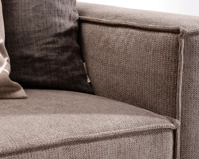 Nærbillede af et sofahjørne med tekstureret, lysebrunt betræk og synlige syninger langs kanterne. Sædet og armlænet har et firkantet, moderne design. En mørkegrå pude og en delvist synlig mønstret pude giver kontrast og komfort.