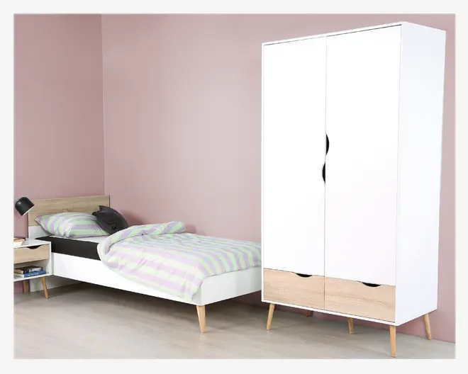 Et moderne soveværelse med en enkeltseng, der har en lys trægavl og hvid ramme, suppleret med skrå træben. Sengen er redt med et stribet dynebetræk i pastelfarverne grøn og lavendel samt en matchende pude og en mørkegrå pyntepude. Ved siden af sengen står et lille natbord med en skuffe og en åben hylde, også med skrå træben, hvorpå der står en sort læselampe og et par bøger. Et stort, hvidt garderobeskab med to låger og to nederste skuffer i lys træfinish står i nærheden, også med skrå træben. Stilen er minimalistisk og skandinavisk inspireret med en blød rosa væg og lyst trægulv.