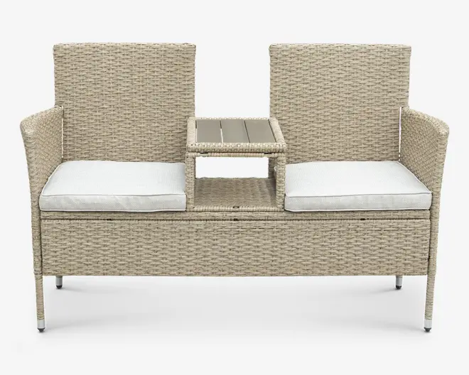 Udendørs bænk med to separate siddepladser, hver med høj ryg og armlæn. Bænken er lavet af lysebeige flettet rattan og har tykke, hvide sædehynder for ekstra komfort. Mellem sæderne er der et hævet centralt bord med en flad overflade, velegnet til at placere drikkevarer eller småting, samt en åben hylde nedenunder til ekstra opbevaring.