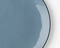 Et nærbillede af en rund tallerken med en glat, blank lyseblå overflade. Tallerkenen har en let ujævn kant og en reflekterende overflade, der fanger lyset og giver et moderne og elegant udtryk.