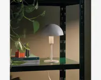 En lille bordlampe med et minimalistisk design, der har en mat hvid, kuppelformet skærm og en slank cylindrisk stander på en rund fod. Lampen står på en grøn hylde ved siden af en stak bøger og en lille dekorationsgenstand. Udtrykket er moderne og enkelt, velegnet til nutidige indretninger.