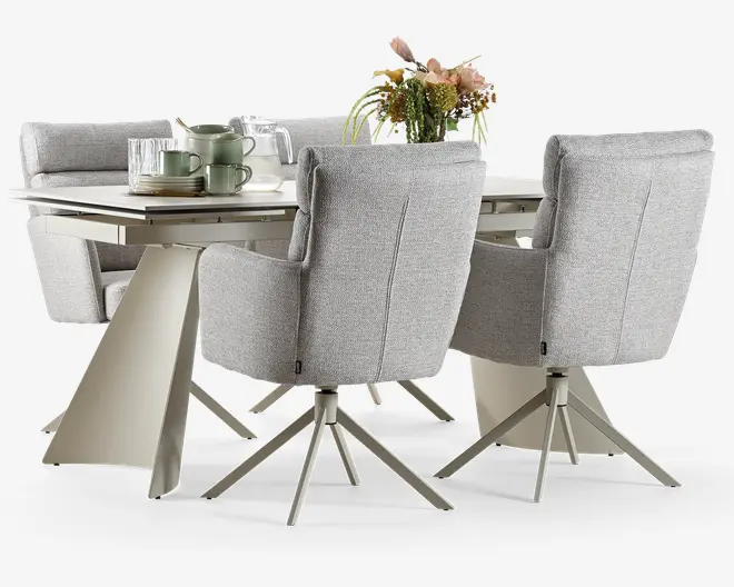 Moderne spisesæt med et rektangulært bord, der har en stilren, skråtstillet metalbase og en glat overflade. Sættet inkluderer polstrede stole og en bænk, alle betrukket med lysegråt stof og diskrete syninger. Stolene har armlæn og drejelige metalben for ekstra komfort og funktionalitet. Bordet er pyntet med en blomsterdekoration samt grønne keramiske tallerkener og glas, hvilket understreger dets moderne udtryk.