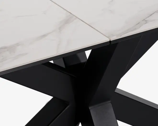 Nærbillede af et bordhjørne med en hvid bordplade i marmorlook med diskrete grå årer og en skrå kant. Bordpladen understøttes af en solid, sort metalbase i krydsform, hvilket giver bordet et moderne og elegant udtryk.