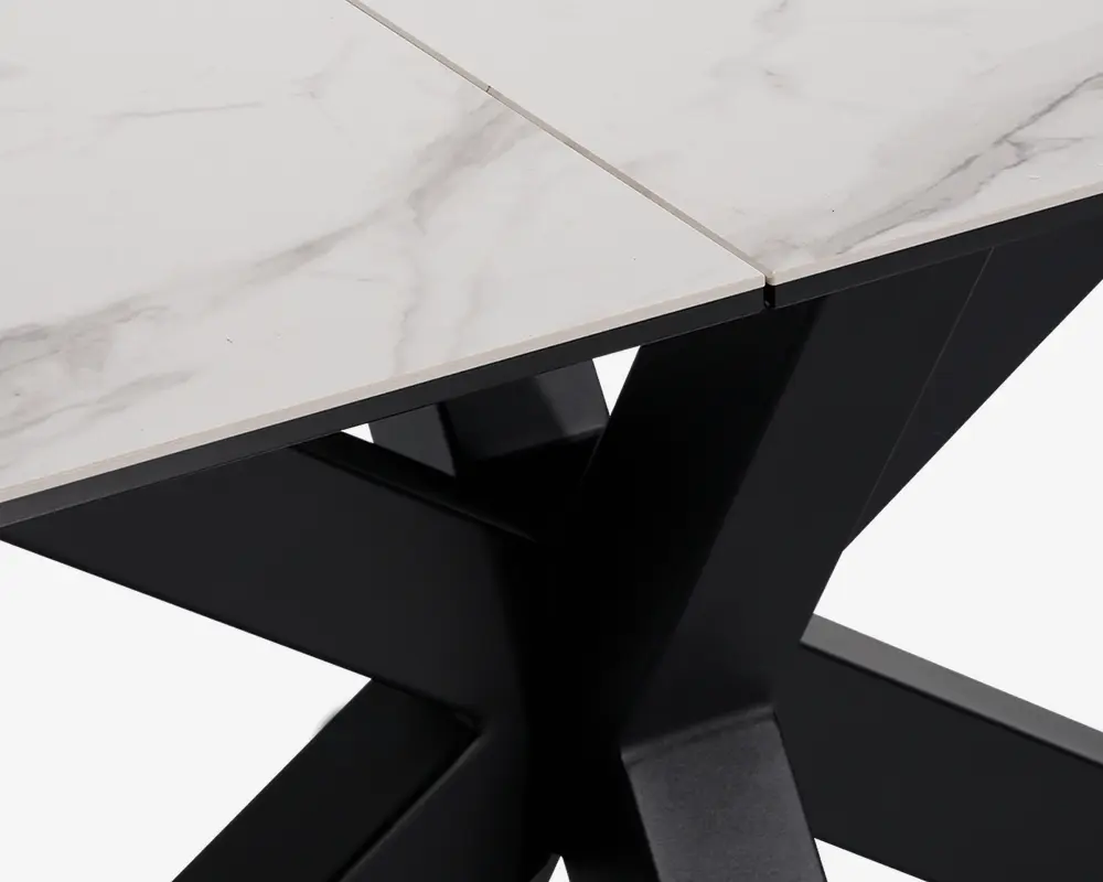 Nærbillede af et bordhjørne med en hvid bordplade i marmorlook med diskrete grå årer og en skrå kant. Bordpladen understøttes af en solid, sort metalbase i krydsform, hvilket giver bordet et moderne og elegant udtryk.