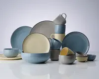 Et udvalg af keramikservice bestående af tallerkener, skåle og krus i matte pastelfarver som blå, grå og creme. Delene har en glat, let blank overflade og et enkelt, moderne design med afrundede kanter. Sættet indeholder både store og små tallerkener, dybe og lave skåle samt stablebare krus, der alle matcher hinanden for et ensartet udtryk på bordet.