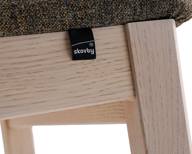 Nærbillede af et møbel, hvor en lys træramme møder en polstret pude med flerfarvet, tekstureret stof. En lille sort stofmærkat med hvid tekst er fastgjort i samlingen mellem træet og puden. Træet har en glat, naturlig overflade med synlige åretegninger.