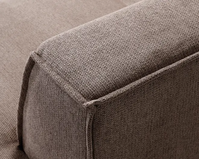 Nærbillede af en sofa-armlæn med tekstureret, taupefarvet stofbetræk. Syningerne er tydelige langs kanterne og giver et pænt og veltilpasset udseende. Stoffet har et vævet look, hvilket giver en blød, men slidstærk overflade. Armlænet er rektangulært med let afrundede hjørner.